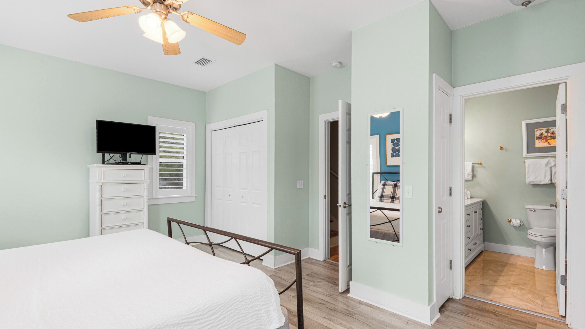 A bedroom with a bed, a TV, a dresser, and a ceiling fan. There is a bathroom visible through an open door.