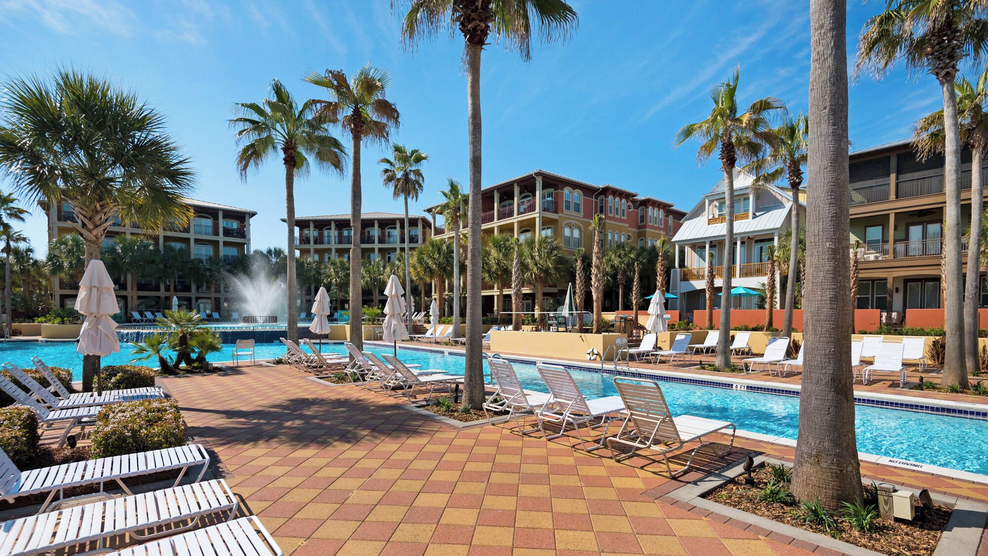 Uma área de piscina de hotel com várias espreguiçadeiras e guarda-sóis, cercada por palmeiras e um céu azul límpido.
