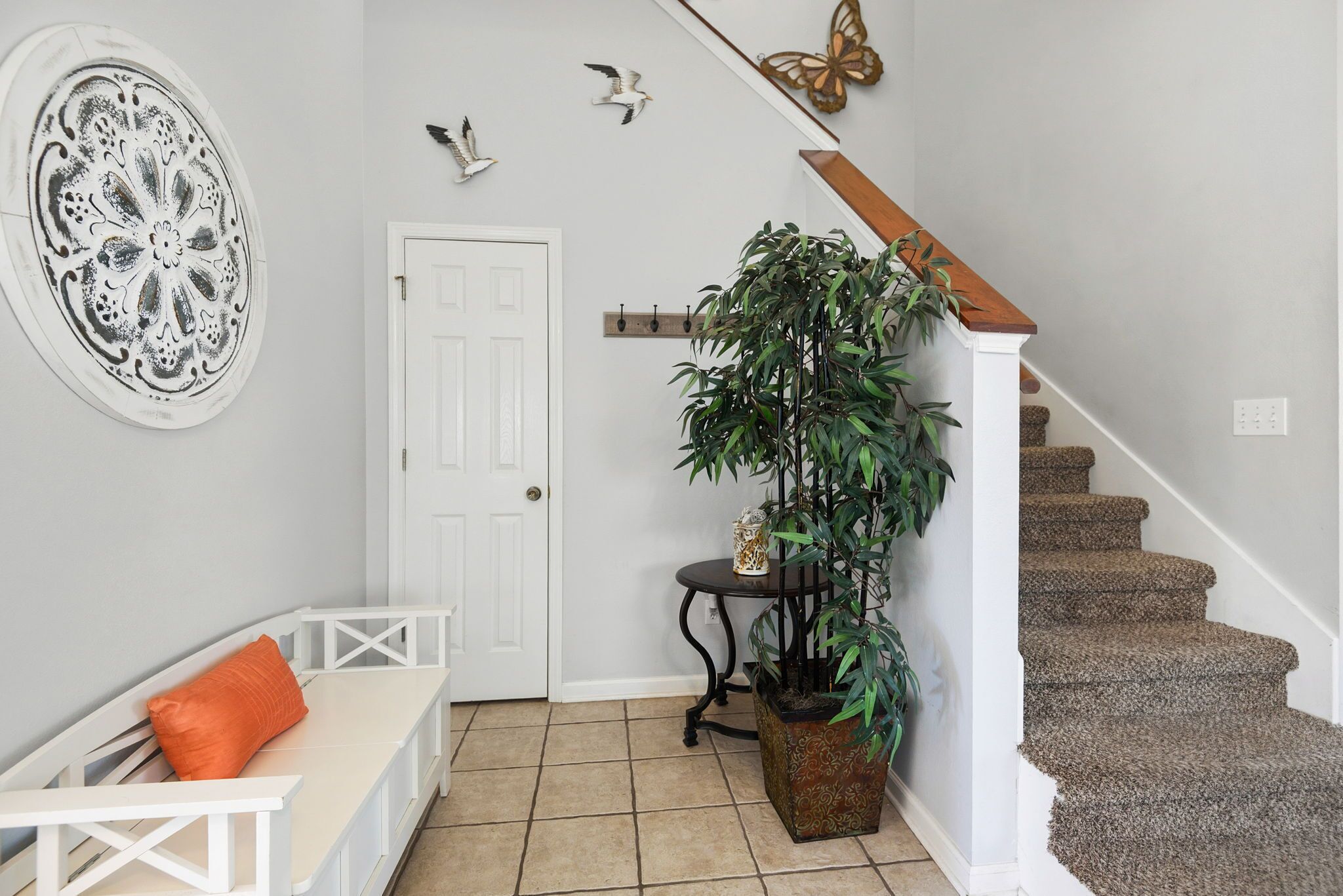 Una escalera con barandilla de madera, una puerta blanca, un banco con cojín naranja, una planta en maceta y decoración de pared que incluye un diseño circular y adornos de pájaros.