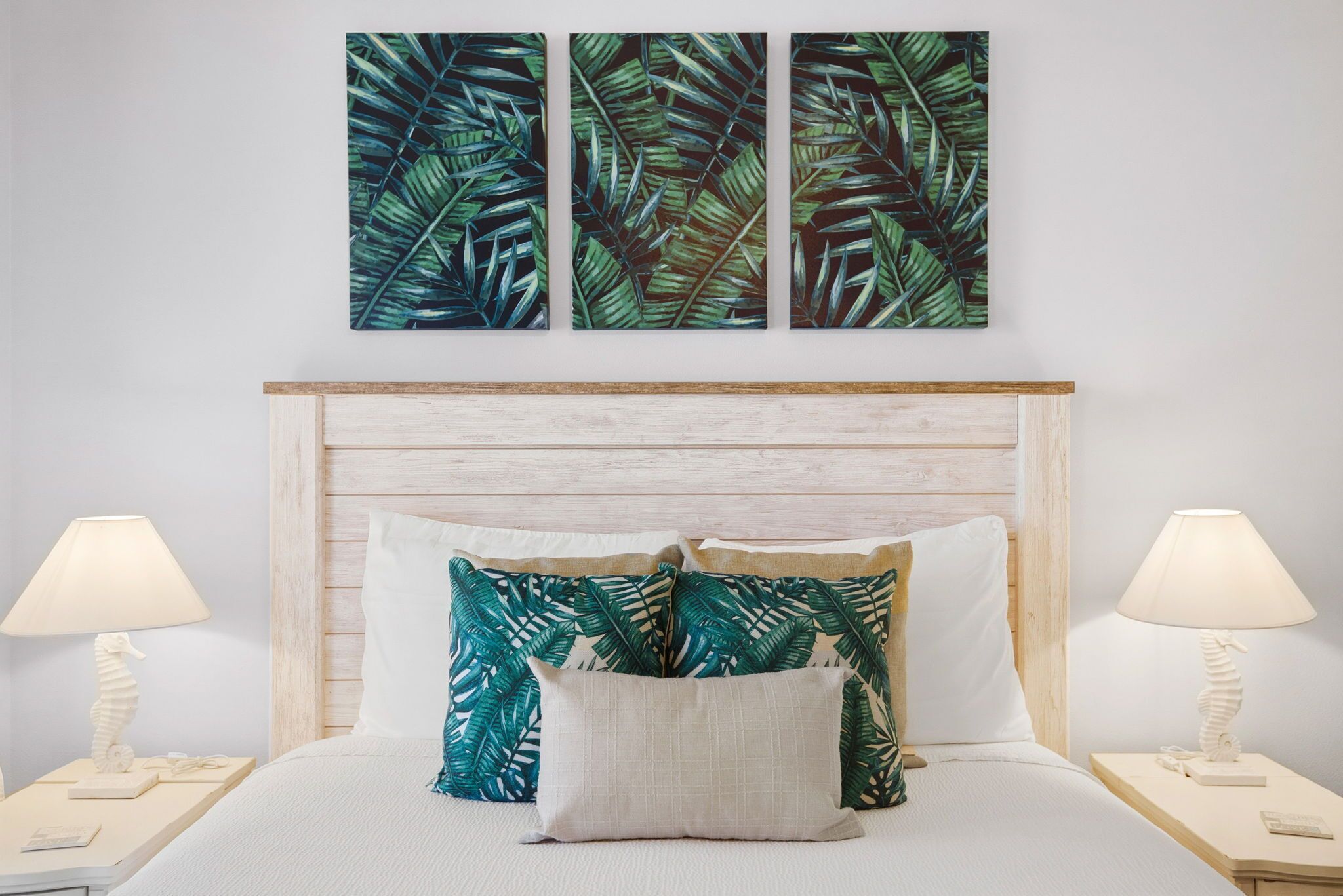 A bedroom with a wooden headboard, a bed with white bedding, two nightstands with lamps, and three wall-mounted leaf-patterned artworks.