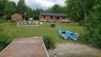 A wooden dock leading to a river, a small cabin, and a larger building with a deck.