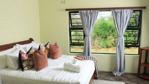 A bed with white bedding, multiple pillows, a bedside table, a window with curtains, and a view of greenery outside. - Perfect 2 Bedroom Farmhouse Close to Bulawayo CBD (Bulawayo)