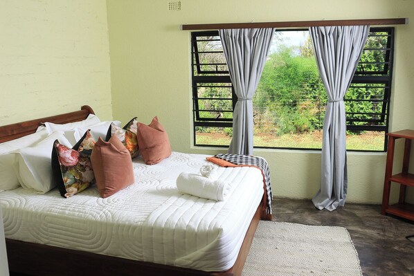 A bed with white bedding, multiple pillows, a bedside table, a window with curtains, and a view of greenery outside.