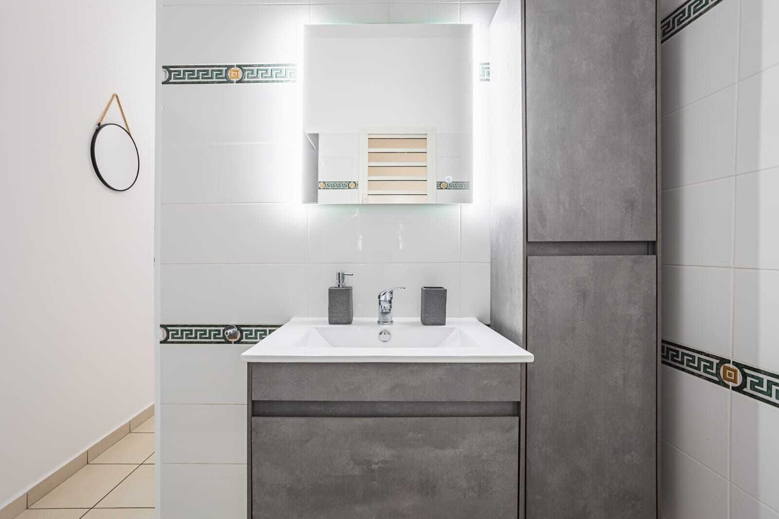 Um banheiro moderno com uma pia branca, dois dispensadores de sabão em tons escuros, um espelho sobre a pia e azulejos decorativos com padrão geométrico.
