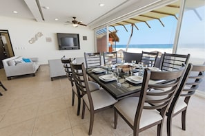 A dining area with a table set for six, a sofa, a TV, and a view of the beach.