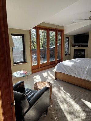 Une chambre à coucher avec un lit, une télévision, une cheminée, une table en verre et une vue sur les arbres à travers de grandes fenêtres.