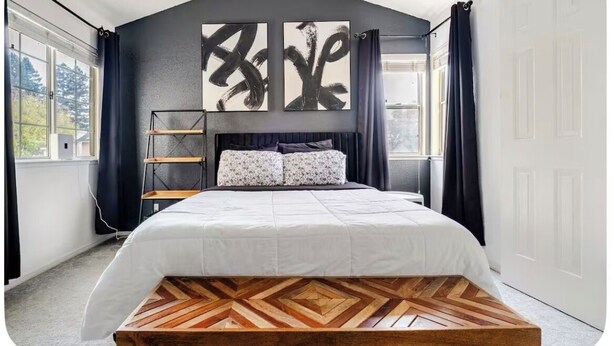 Un dormitorio con una cama, una plataforma de madera, un cuadro abstracto negro y una ventana con cortinas.