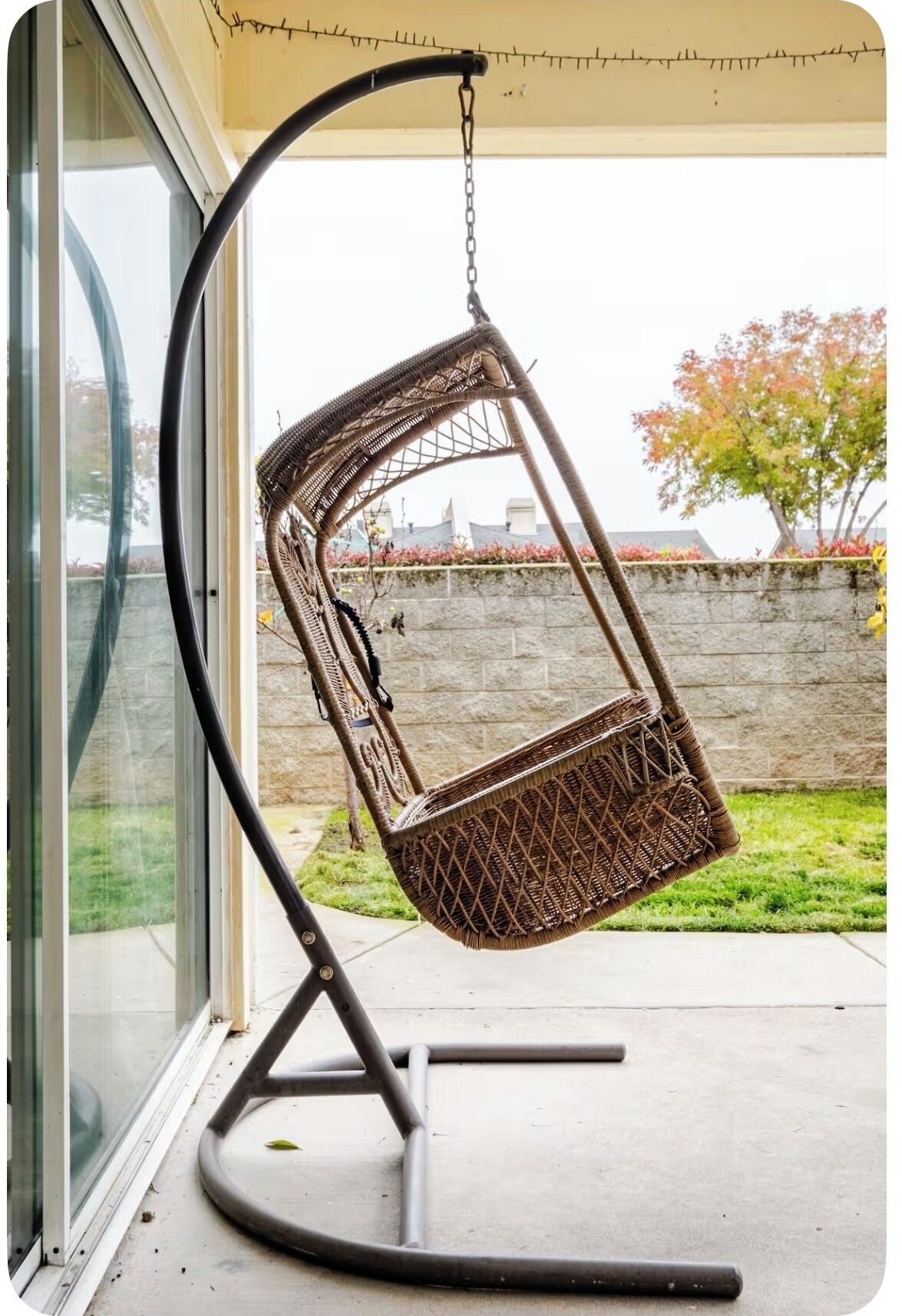 Un fauteuil suspendu en osier, fixé à une structure métallique, placé devant une porte vitrée.