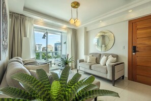 A modern living room with a sofa, a coffee table, a large plant, a round mirror, and a view of the city through large windows. - Luxury 2BR in Bella Vista | Pool & Comfort for 8 (Santo Domingo)