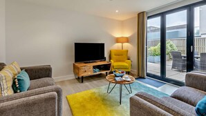 A modern living room with a flat-screen TV, a yellow armchair, and a dining area with wicker furniture. - Mist - Luxury Cottages (Woolacombe)