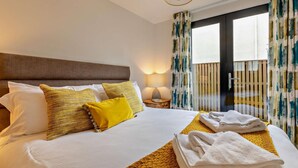 A hotel room with a bed, pillows, a nightstand, a lamp, and a view of the outside. - Mist - Luxury Cottages (Woolacombe)