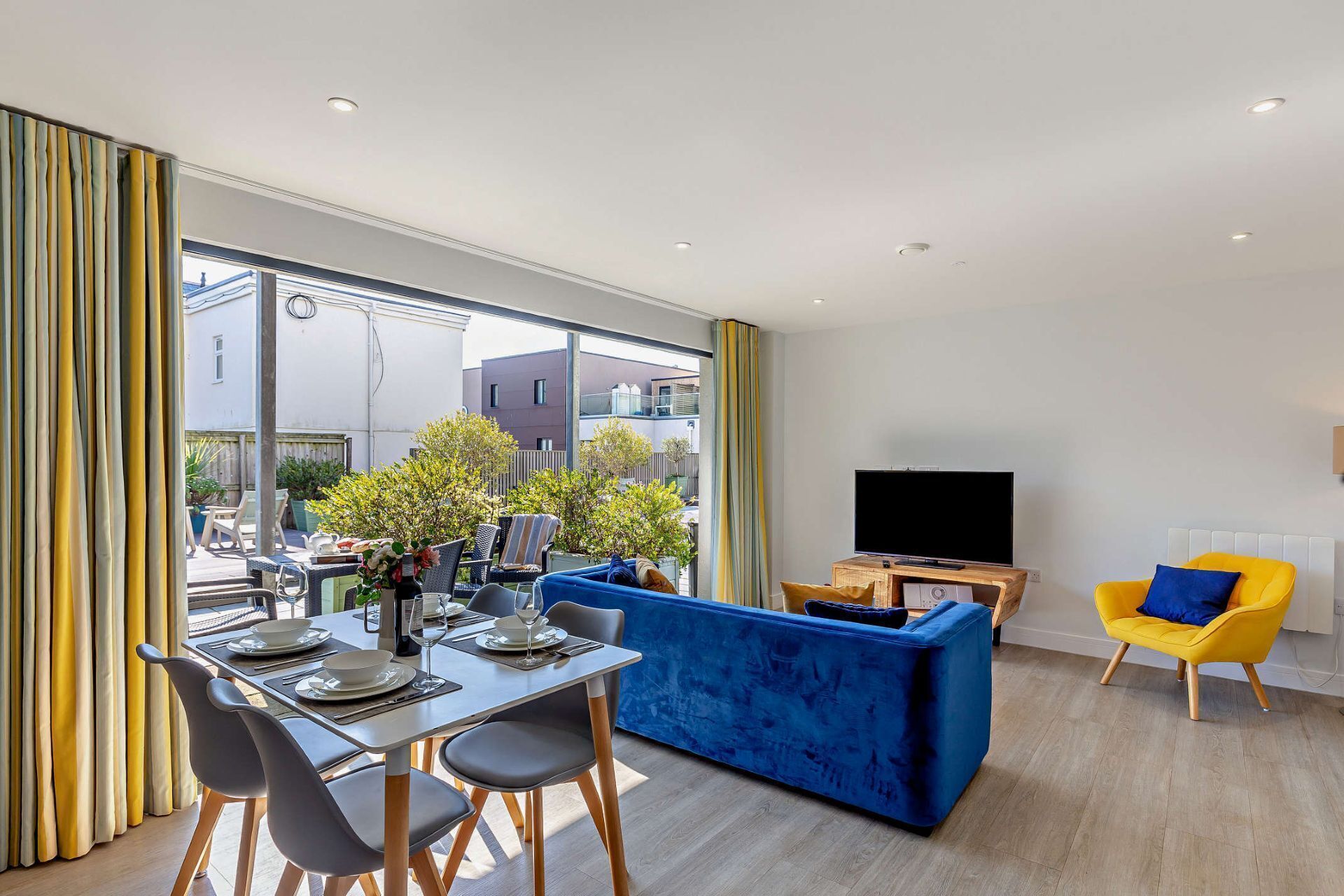 Uma sala de estar moderna com área de jantar, um sofá azul, uma poltrona amarela e uma televisão de tela plana.