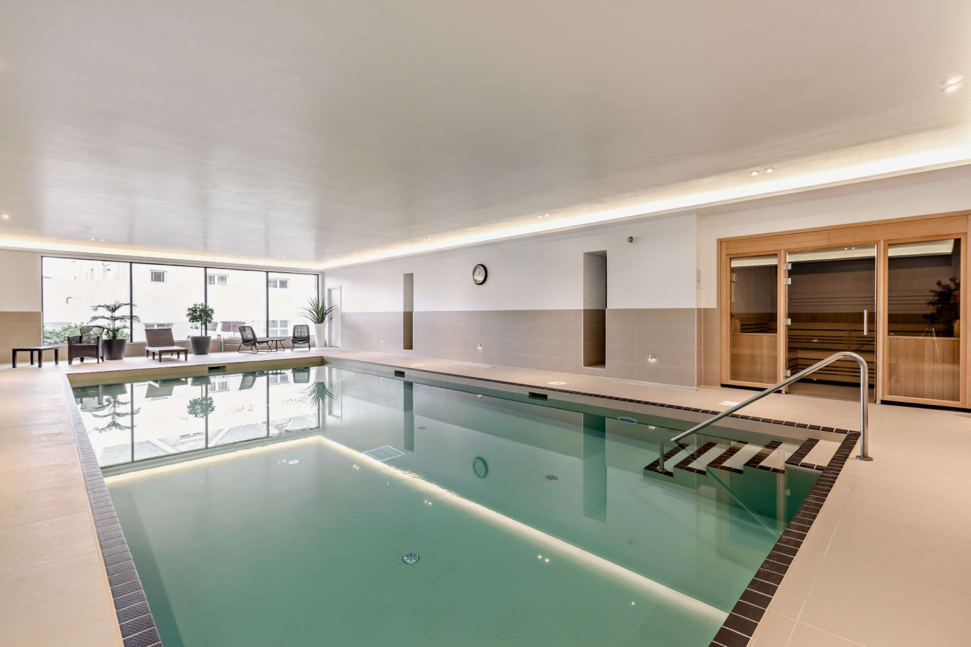 An indoor swimming pool with a clear view of the pool area, a clock on the wall, and a seating area with chairs and tables.