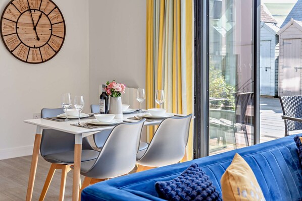 A dining area with a table set for four, chairs, a clock, and a view through a large window.
