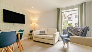 A living room with a sofa, chairs, a flat-screen TV, a lamp, and a view of buildings outside. - Waveside - Luxury Cottages (Woolacombe)