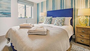 A neatly made bed with pillows and a folded towel, a bedside table with a lamp, a window with blinds, and a striped wall. - Waveside - Luxury Cottages (Woolacombe)