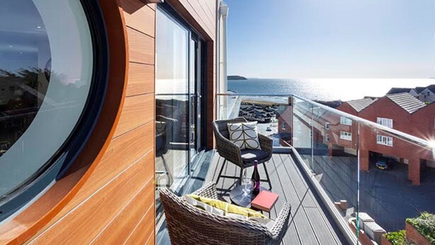 A balcony with a wicker chair, a small table, and a balcony railings. - Reefside - Luxury Cottages (Woolacombe)