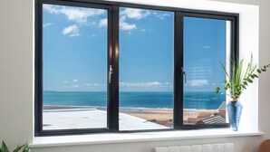 A window with a view of the sea, a white radiator below the window, and a plant in a vase. - Reefside - Luxury Cottages (Woolacombe)