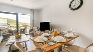 A dining area with a wooden table set for four, a television, and a view of the outdoors. - Wavemist -Luxury Cottages (Woolacombe)