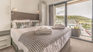 A hotel room with a bed, a bedside table, a lamp, and a large window with a view of a town. - Wavemist -Luxury Cottages (Woolacombe)