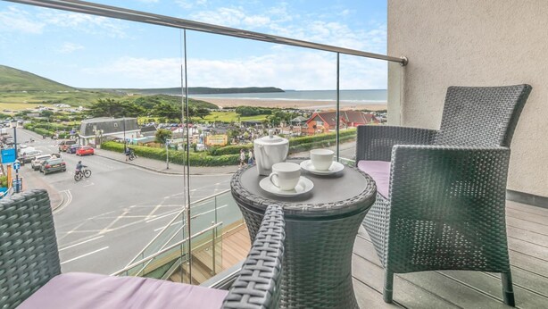 A balcony with wicker furniture, a table with cups and a teapot, and a view of a coastal town. - Wavemist -Luxury Cottages (Woolacombe)