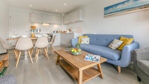 A modern living room with a blue sofa, wooden coffee table, and a dining area with chairs and a table. - Wavemist -Luxury Cottages (Woolacombe)