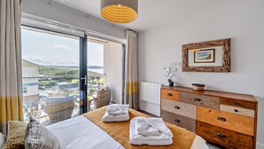 A hotel room with a bed, a wooden dresser, a framed picture, and a view of the landscape. - Whitehaven - Luxury Cottages (Woolacombe)