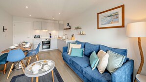A modern living room with a blue sofa, a dining table with chairs, a kitchen area with cabinets and appliances, and a framed picture on the wall. - Whitehaven - Luxury Cottages (Woolacombe)