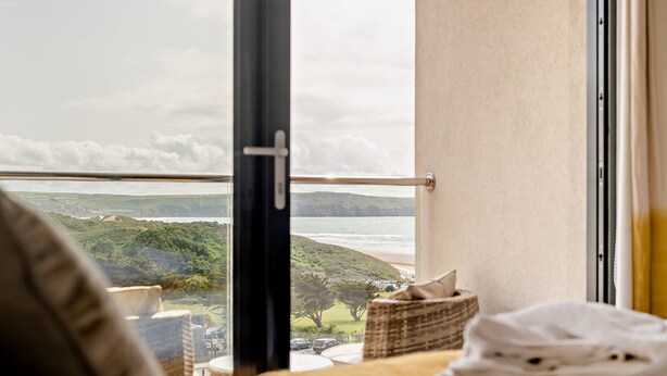 A room with a large window overlooking a coastal landscape, a bed, a bedside table with a folded towel, and a wicker basket. - Whitehaven - Luxury Cottages (Woolacombe)