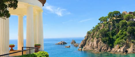 A seaside view with a white pavilion, clear blue sky, rocky coastline, and lush greenery.