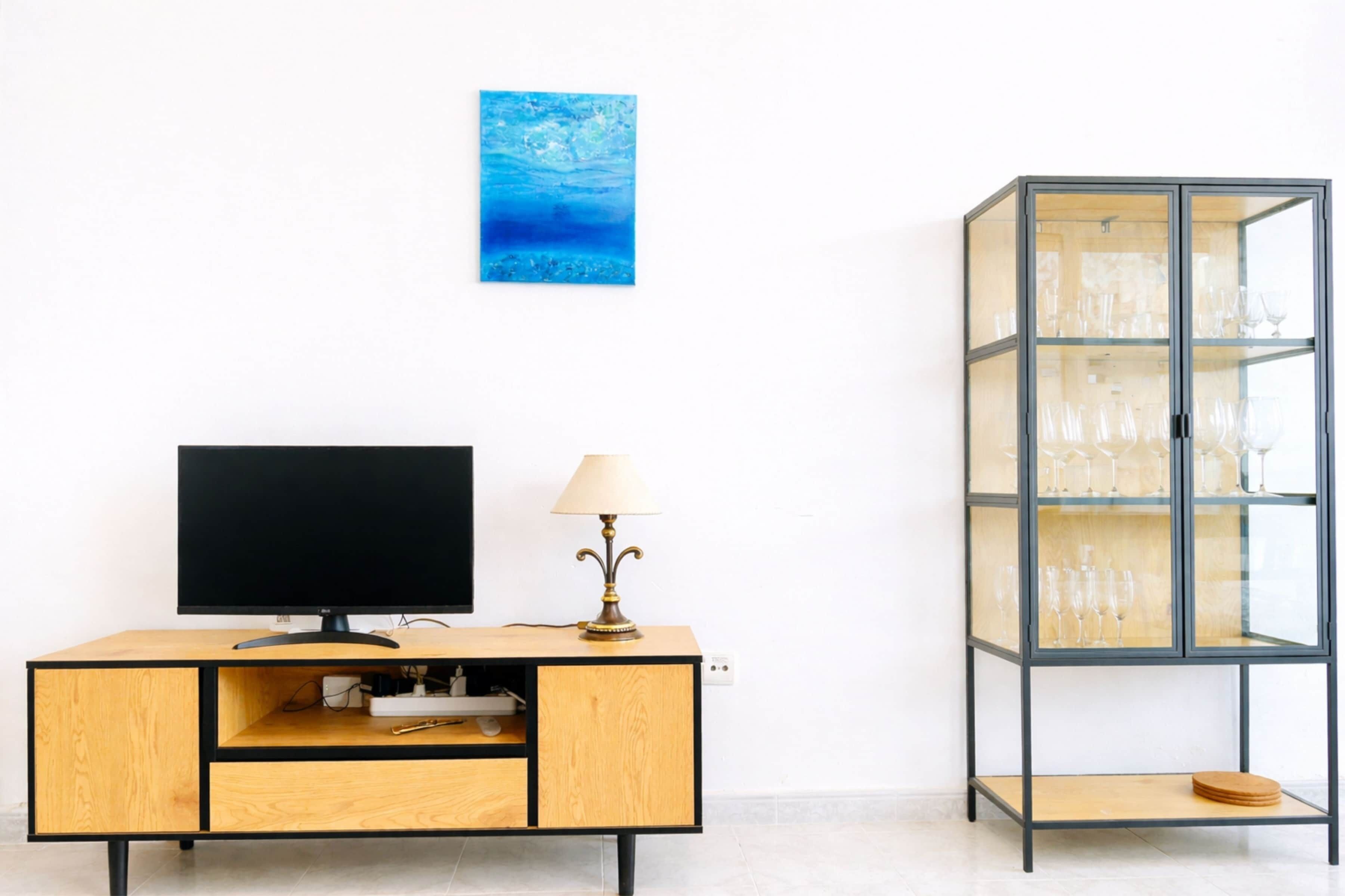 A modern living room with a flat-screen TV, a wooden cabinet, a glass display cabinet, and a blue artwork on the wall.