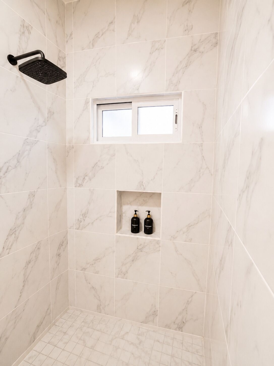 A bathroom with marble tiles, a showerhead, a window, and two bottles on a shelf.
