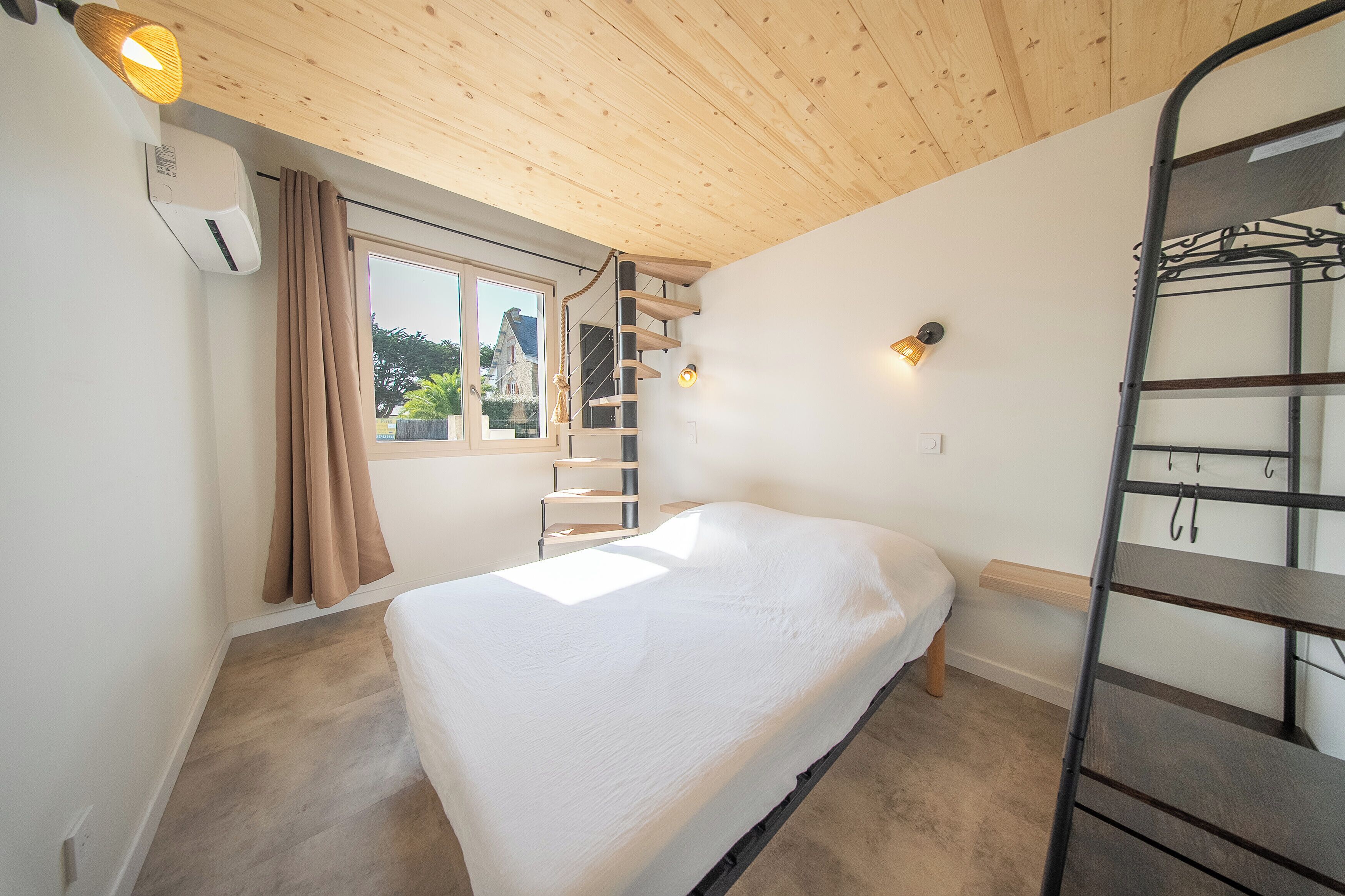A single-bed room with a wooden ceiling, a window with curtains, a ladder, and a wall-mounted shelf.