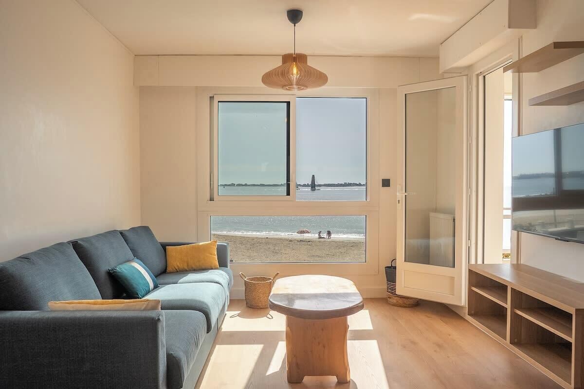 A modern living room with a sofa, a coffee table, and a large window overlooking a beach.