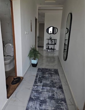 A hallway with a marble floor, a potted plant, a rug, a mirror, and a door leading to a bathroom.