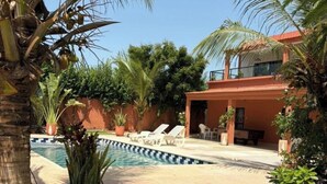 Una piscina con sillones de descanso, una palmera y un edificio con techo rojo.