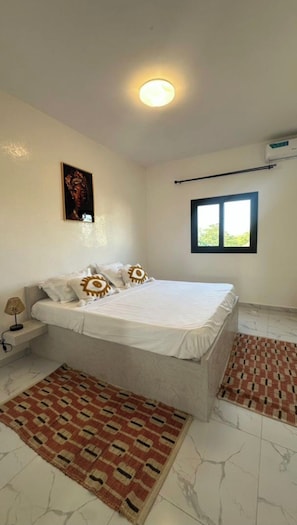 A bedroom with a bed, a nightstand, a lamp, a window, and a wall-mounted air conditioner.