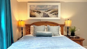 A neatly made bed with a headboard, two bedside lamps, a framed painting above the bed, and a window with curtains.