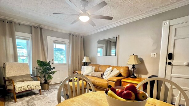A cozy living room with a wooden dining table, a sofa, a ceiling fan, and a view of the outdoors through a window.