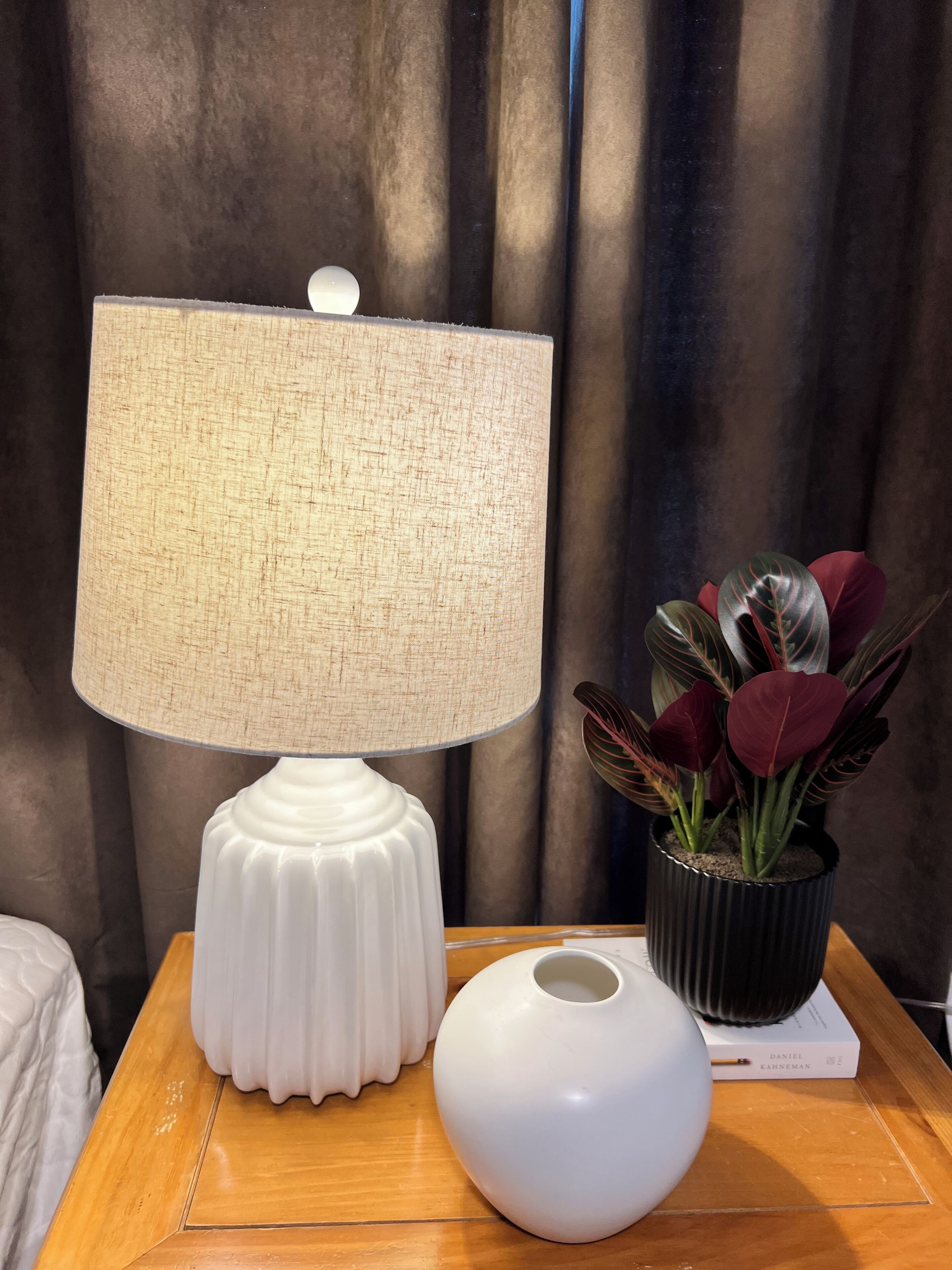 Una lámpara de mesa blanca con pantalla texturizada, un florero blanco y una planta en maceta con hojas rojas y verdes sobre una mesita de noche de madera.