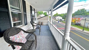 A porch with wicker chairs and a table, overlooking a street with houses and a pedestrian crossing.
