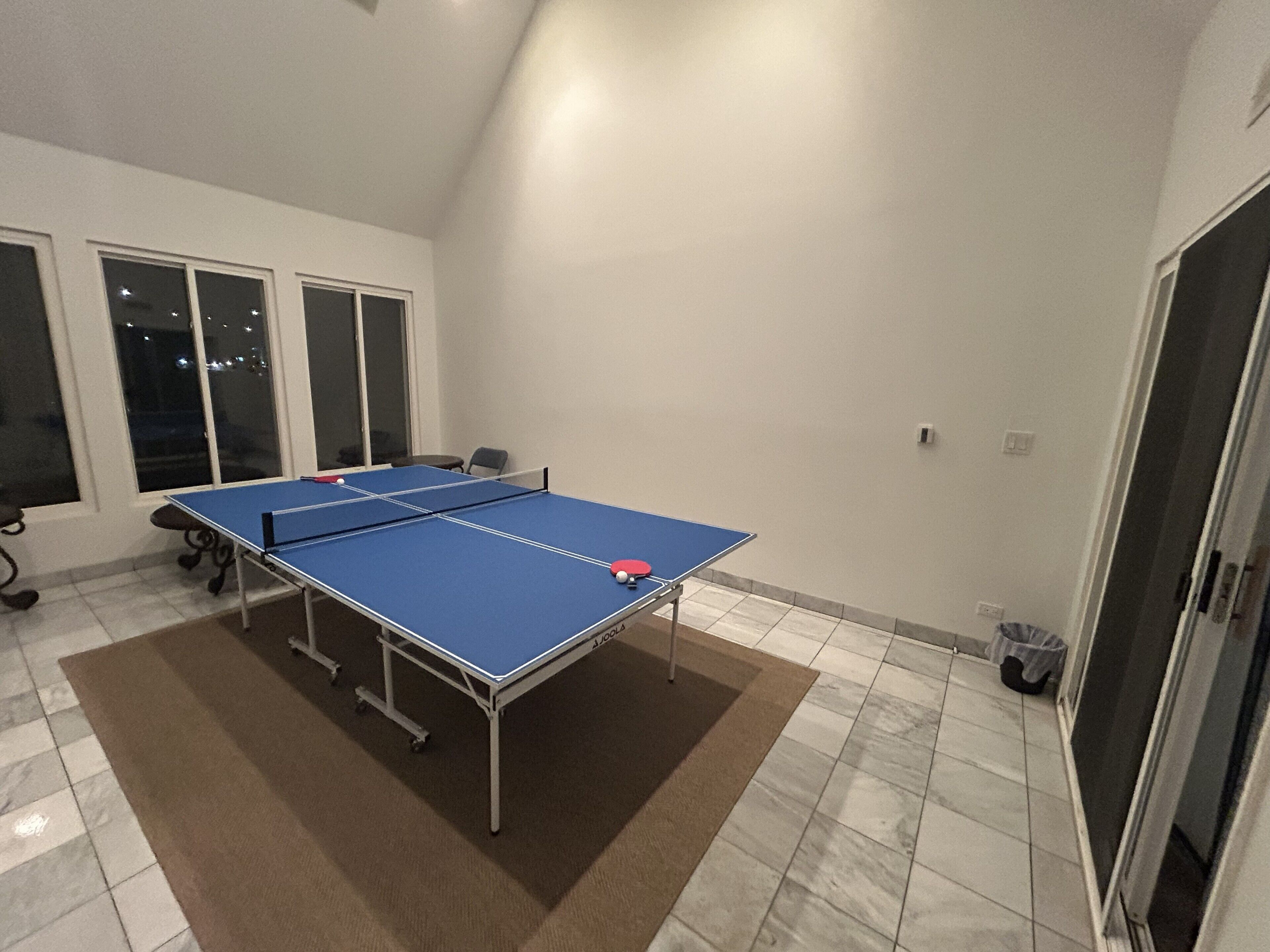 A ping pong table in a room with a tiled floor, large windows, and a door.