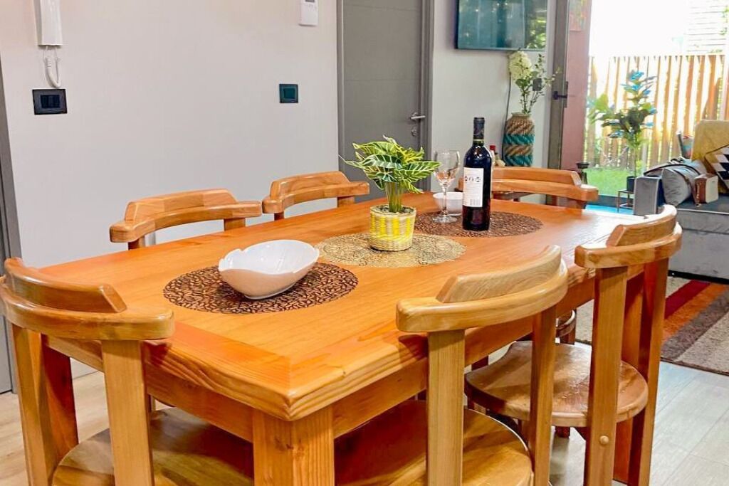 A wooden dining table with six chairs, a white bowl, a bottle of wine, and a vase with green plants.