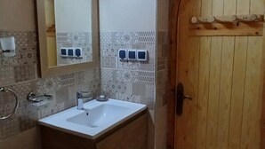 A bathroom with a wooden door, a sink, a mirror, and a wall-mounted light fixture. - La Colline Verte Dar Ait El Abass  (Skoura)