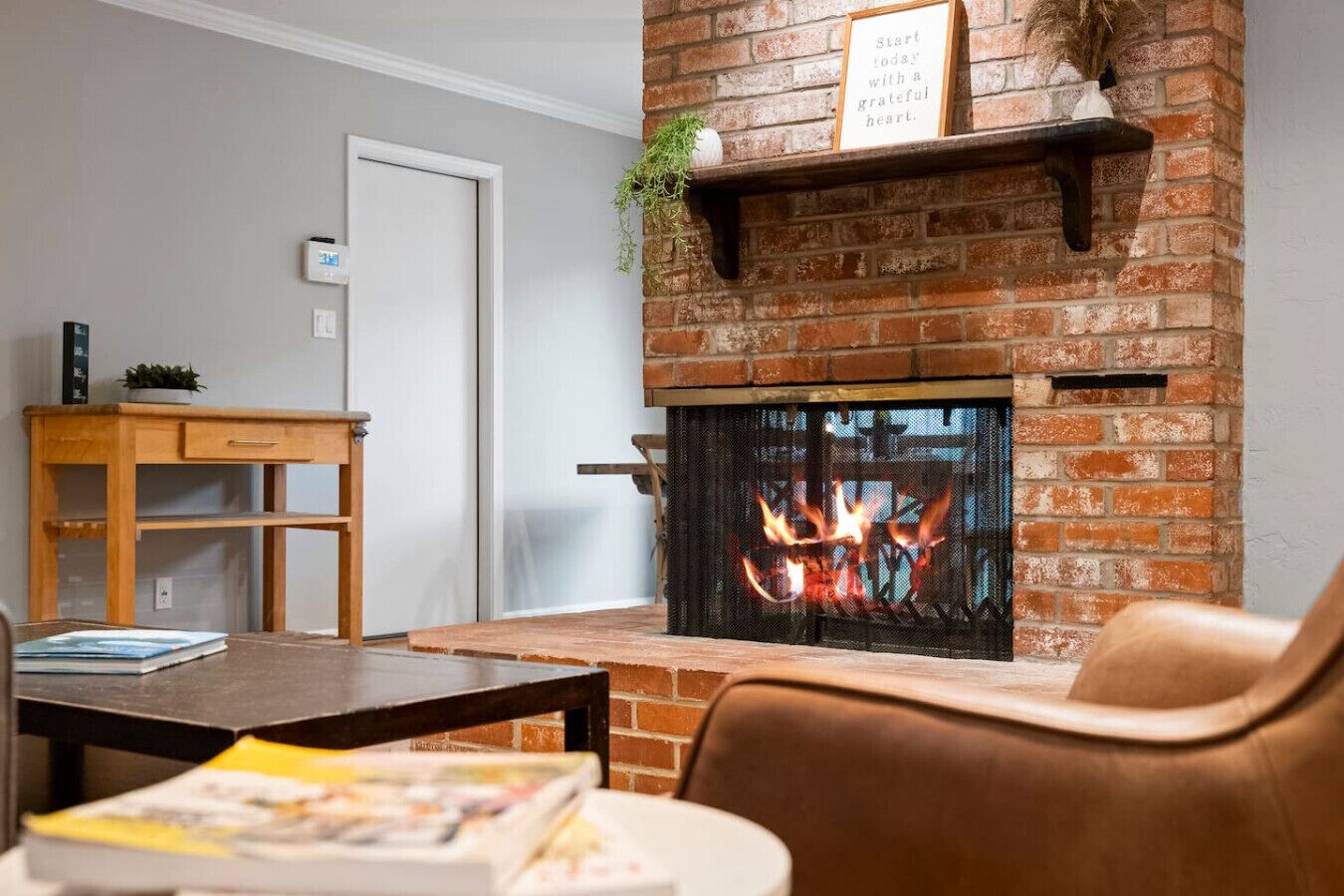 A living room with a brick fireplace, a wooden table, a chair, and a small plant on the mantel.
