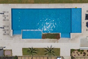 Eine Vogelperspektive auf einen rechteckigen Swimmingpool mit einer umlaufenden Liegefläche, Liegestühlen und einem kleinen Gartenbereich mit Pflanzen.