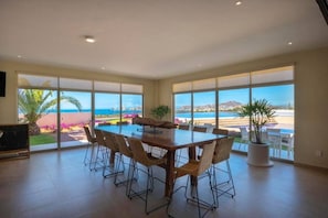 A spacious dining area with a large table and chairs, large windows with a view of the beach and ocean, and a potted plant. - The Arch View — Vista Vela (Cabo San Lucas)