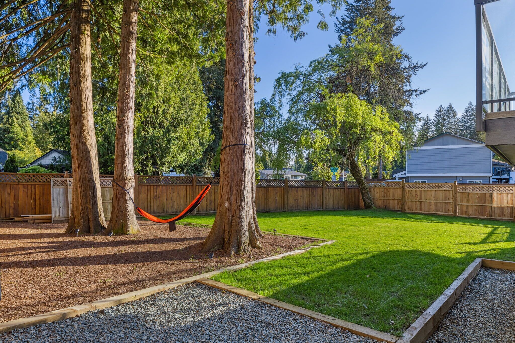 A backyard with a hammock, trees, a wooden fence, and a well-maintained lawn.