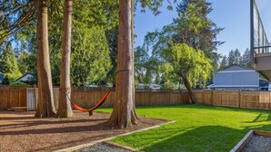 Un patio trasero con una hamaca, árboles, una cerca de madera y un césped bien cuidado.