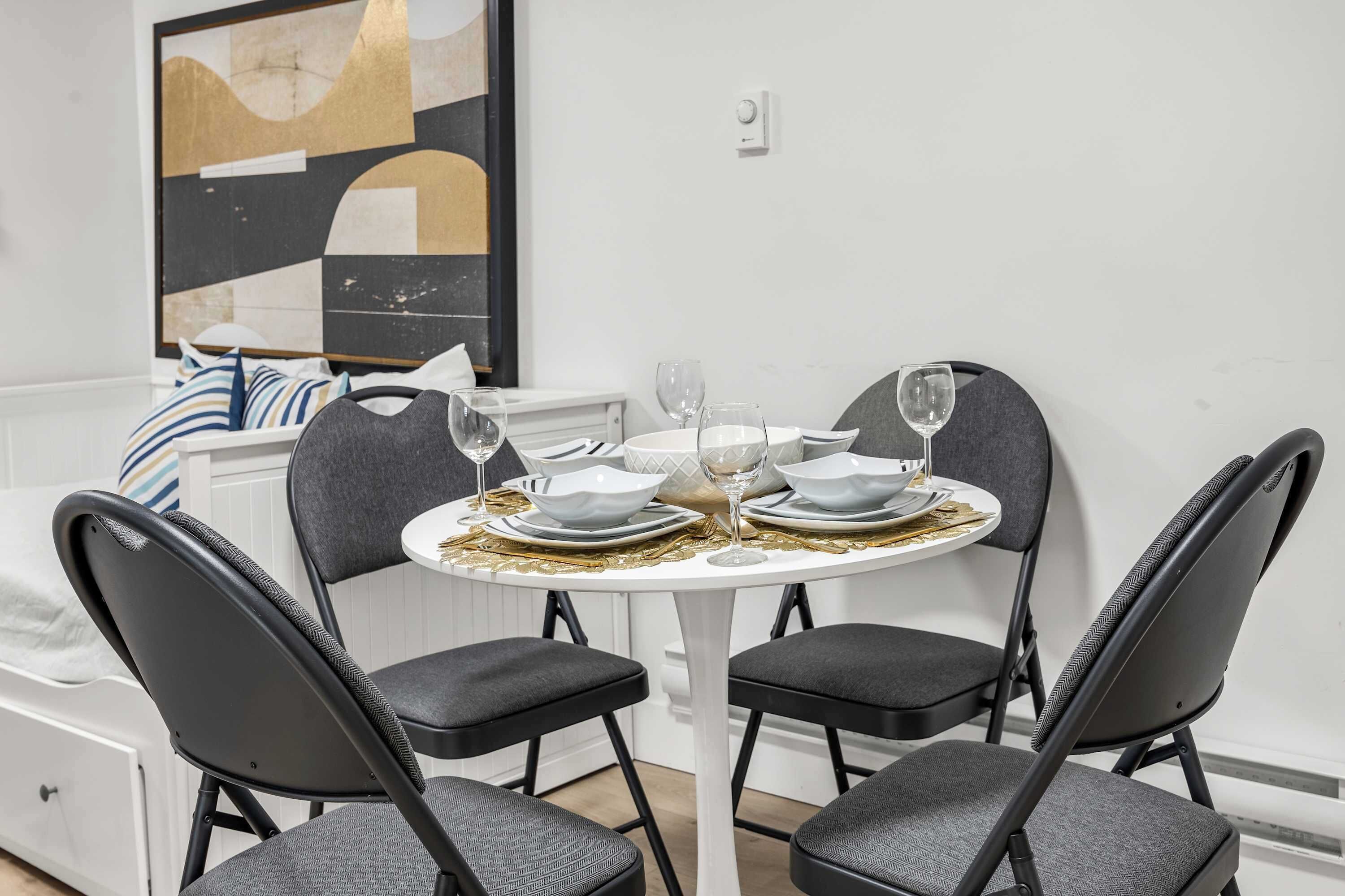 A dining area with a round table, four chairs, and a white wall with a painting.